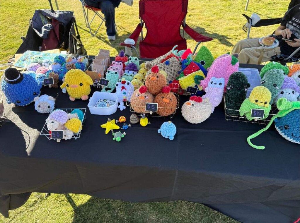 Close-up of a vendor booth at Family Fun Day showcasing handmade crochet plush toys arranged in wire baskets on a black tablecloth. The colorful crafts include animals and decorative figures, each with small price tags. The table is set outdoors on grass, adding to the festive community atmosphere.