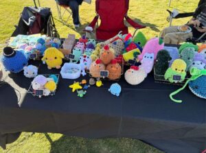 Close-up of a vendor booth at Family Fun Day showcasing handmade crochet plush toys arranged in wire baskets on a black tablecloth. The colorful crafts include animals and decorative figures, each with small price tags. The table is set outdoors on grass, adding to the festive community atmosphere.