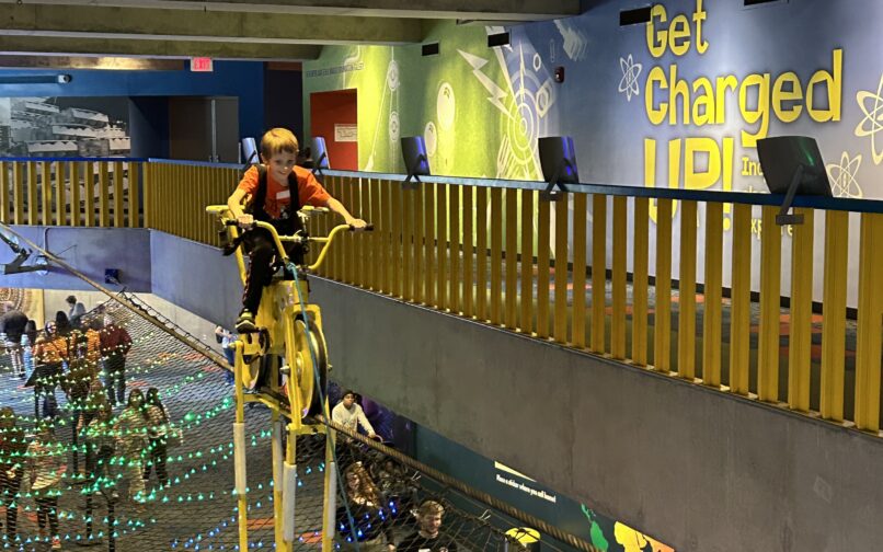 A child rides a yellow suspended bicycle on a high‑wire exhibit above a museum walkway, while visitors walk below through a netted area. Colorful lights and a “Get Charged Up!” science‑themed wall display decorate the space.
