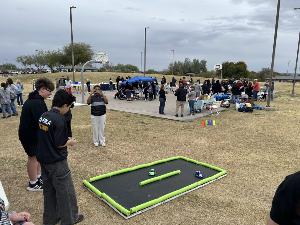 Outdoor event at a park with groups of people gathered around tables and activity stations near a basketball court. In the foreground, two individuals stand near a small foam-bordered racetrack on the grass, with toy cars positioned inside it. A person in the mid-ground is taking a photo, and tents, cones, and various activity setups are visible throughout the area under cloudy skies.