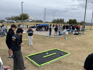 Outdoor event at a park with groups of people gathered around tables and activity stations near a basketball court. In the foreground, two individuals stand near a small foam-bordered racetrack on the grass, with toy cars positioned inside it. A person in the mid-ground is taking a photo, and tents, cones, and various activity setups are visible throughout the area under cloudy skies.